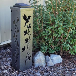 May include: A metallic gray solar-powered garden light. The rectangular light has cut-out floral and hummingbird designs. The top has a solar panel. The light is next to green bushes and rocks.