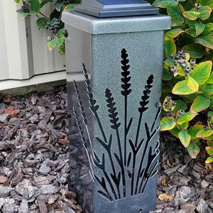 May include: A rectangular solar-powered garden light with a textured silver-gray finish. The front panel features a laser-cut design of tall, slender stalks of lavender. A black solar panel is mounted on the flat top of the light fixture.