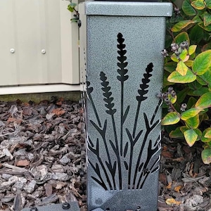 May include: A rectangular, silver-toned metal outdoor light fixture with a textured, speckled finish. The front panel features a laser-cut design of lavender stalks. Two additional matching metal plates with black fasteners are positioned in front.