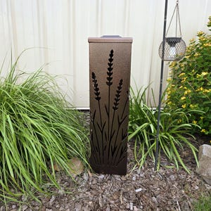 May include: A tall, rectangular, bronze-colored metal garden light featuring laser-cut silhouettes of lavender stalks on its front panel. The fixture has a flat top and a textured finish, standing upright among garden plants and mulch.