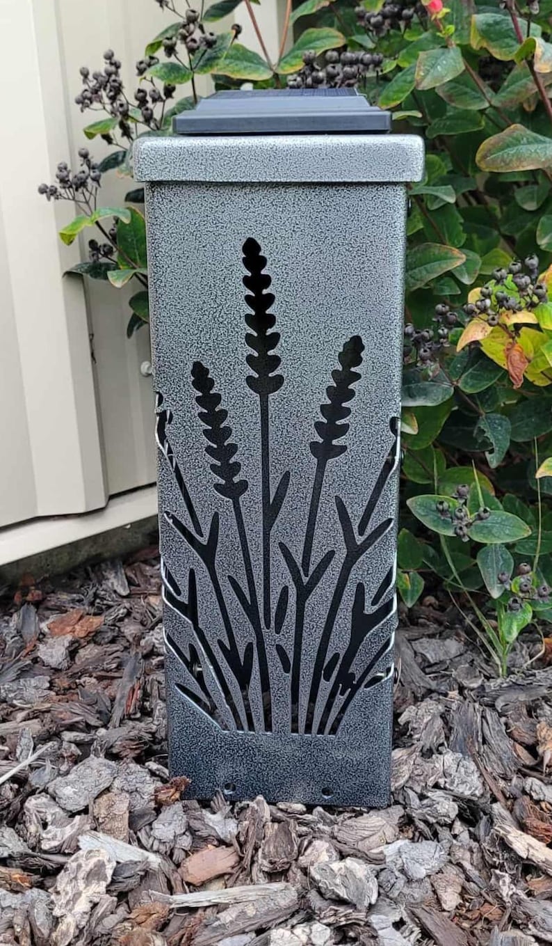 May include: A rectangular solar garden light with a textured silver-gray finish. The front panel features a laser-cut design of three stalks of lavender or wheat. The top of the light is flat and black, and the structure has a sturdy, upright form.