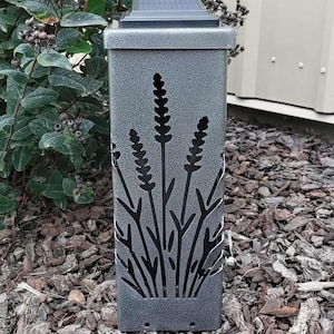 May include: A rectangular solar-powered garden light with a textured gray finish. The metal exterior features a laser-cut design of lavender stalks. A black solar panel is mounted on the flat top of the fixture, which stands upright.