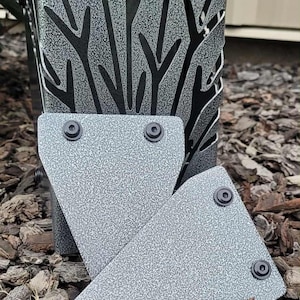 May include: A gray metal object with a textured finish and a cutout design of tall grass or wheat stalks. Two triangular metal plates with black circular fasteners are positioned in front of the main structure, featuring the same gray textured finish.