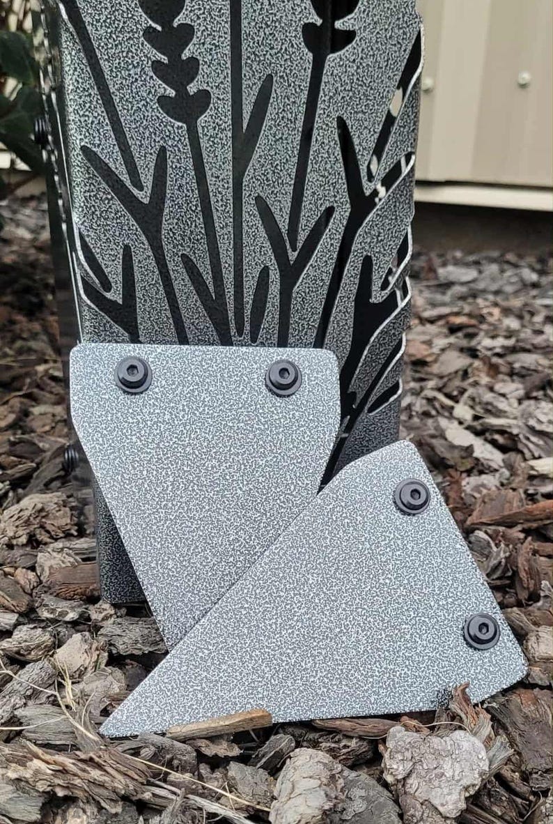 May include: A gray metal object with a textured finish and a cutout design of tall grass or wheat stalks. Two triangular metal plates with black circular fasteners are positioned in front of the main structure, featuring the same gray textured finish.