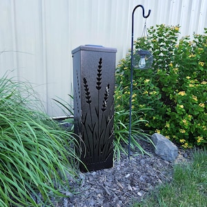 May include: A tall, rectangular, dark bronze metal garden light featuring laser-cut silhouettes of lavender stalks on its front panel. The structure has a textured finish and a flat top, standing upright among garden plants and dark mulch.