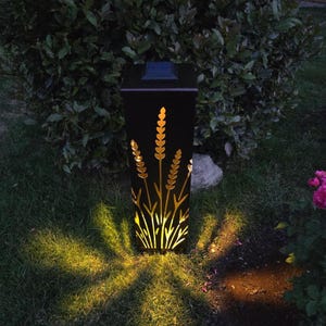 May include: A black square solar garden light with a laser-cut design of wheat stalks on its front panel. The light emits a warm yellow glow through the cutouts, casting a starburst pattern onto the grass below. A small solar panel sits on top.