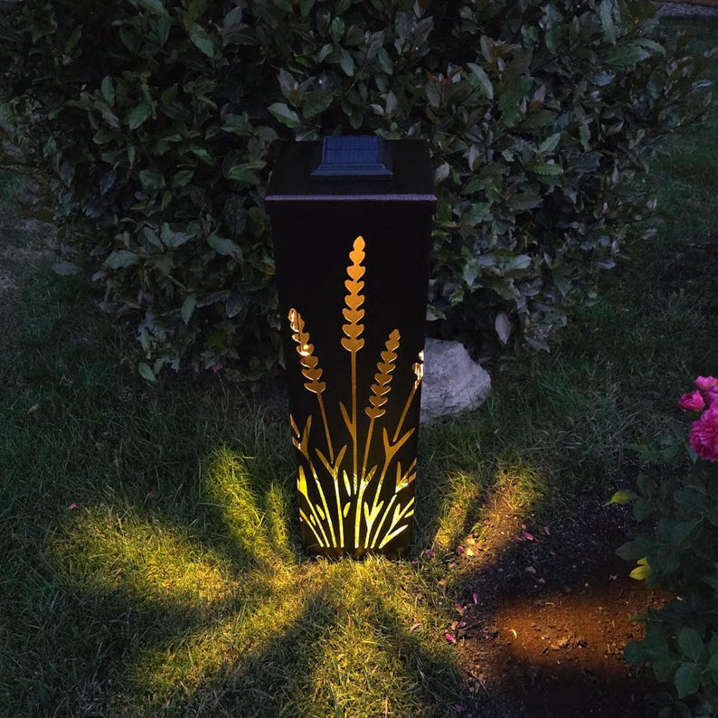 May include: A black square solar garden light with a laser-cut design of wheat stalks on its front panel. The light emits a warm yellow glow through the cutouts, casting a starburst pattern onto the grass below. A small solar panel sits on top.
