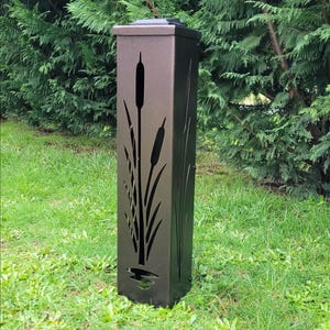 May include: A tall, rectangular, dark brown metal outdoor post with a cattail design cut out of the front. The post is set in a grassy area with green foliage in the background.
