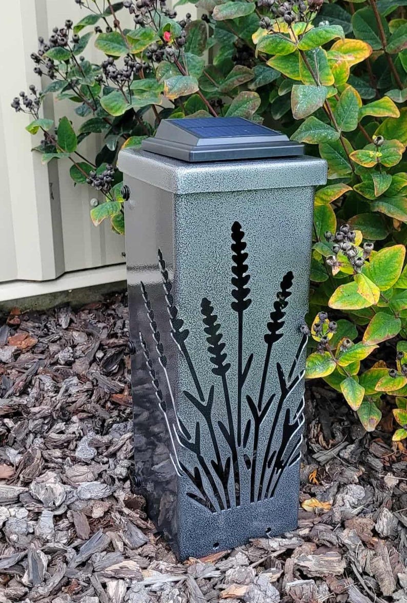 May include: A rectangular solar-powered garden light with a textured silver-gray finish. The front panel features a laser-cut design of tall, slender stalks of lavender. A black solar panel is mounted on the flat top of the light fixture.