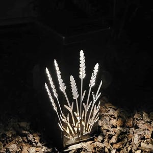 May include: A square, black metal outdoor lantern featuring intricate, laser-cut silhouettes of wheat stalks or tall grasses. The internal light source illuminates the cutouts, creating a bright, glowing pattern against the dark, solid frame.