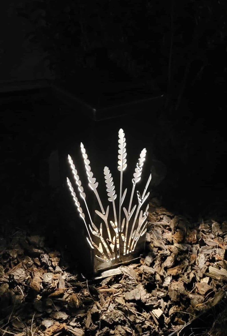 May include: A square, black metal outdoor lantern featuring intricate, laser-cut silhouettes of wheat stalks or tall grasses. The internal light source illuminates the cutouts, creating a bright, glowing pattern against the dark, solid frame.