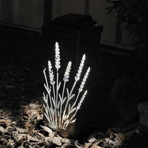 May include: A rectangular black outdoor solar light fixture featuring a laser-cut design of lavender stalks on its front panel. The light emits a bright white glow through the cutouts, illuminating the floral pattern against the dark metal frame.