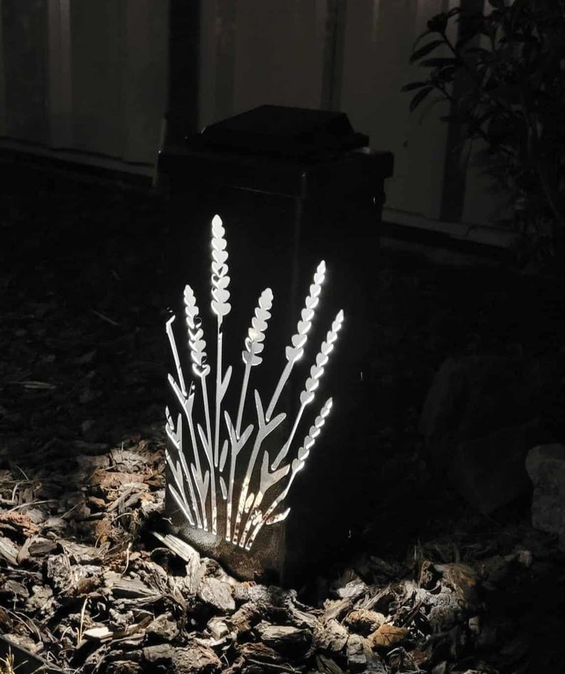May include: A rectangular black outdoor solar light fixture featuring a laser-cut design of lavender stalks on its front panel. The light emits a bright white glow through the cutouts, illuminating the floral pattern against the dark metal frame.