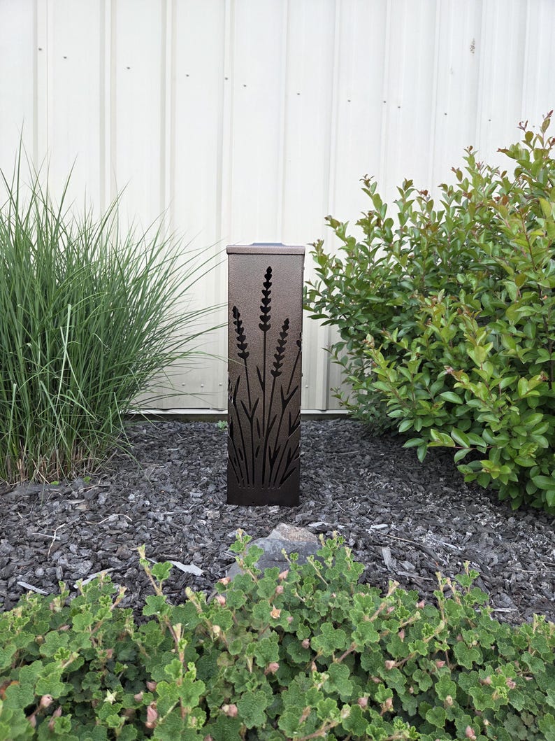 May include: A rectangular dark brown metal garden light featuring a laser-cut design of lavender stalks and foliage. The vertical fixture has a flat top and a textured finish, with cutouts that allow light to pass through the metal structure.