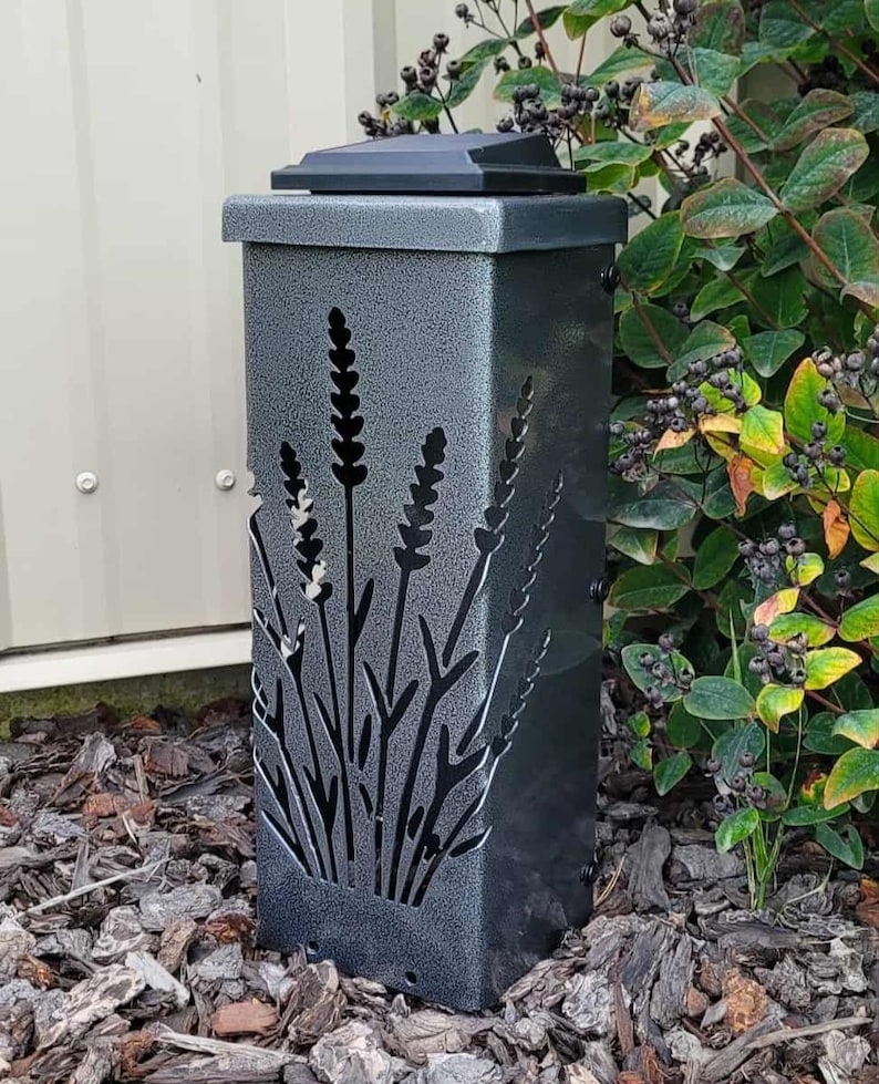 May include: A rectangular, metallic gray solar light fixture with a textured, hammered finish. The front panel features a laser-cut design of lavender stalks, allowing light to pass through the cutouts. It has a flat, black solar panel top.