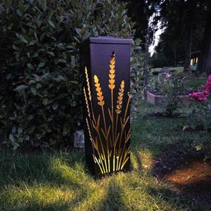 May include: A tall, rectangular black metal outdoor lantern featuring laser-cut designs of wheat stalks or lavender stems. The structure emits a warm, golden light from within, illuminating the cutouts against the dark, textured metal surface.