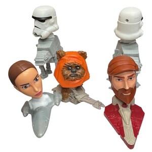 May include: A collection of Star Wars character figurines, including a Stormtrooper, an Ewok, and other characters. The figurines are in various poses and colors, with detailed facial features and costumes. The figures are made of plastic.