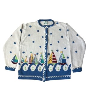 May include: White button-up cardigan sweater with a nautical theme. The sweater features blue star and sailboat designs. The bottom hem has a blue wave pattern with anchor details. The collar and button trim are also blue.