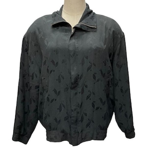 May include: A black long-sleeve jacket featuring a tonal leaf pattern throughout the fabric. The jacket includes a high collar, a full-length front zipper closure, and ribbed cuffs at the wrists and hemline, providing a structured fit.