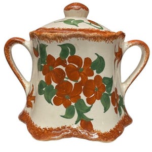 May include: A ceramic sugar bowl with a lid. It has a white base decorated with orange flowers and green leaves. The rim and handle are trimmed with a brown, textured border. The sugar bowl has two handles.