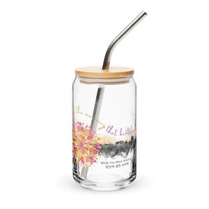 Puede incluir: Un vaso transparente con tapa de bambú y pajita de metal plateado. La taza presenta un diseño floral en tonos rosa, naranja y amarillo, con el texto "the morning that banished" y "While You Were Sleeping" en inglés y coreano.
