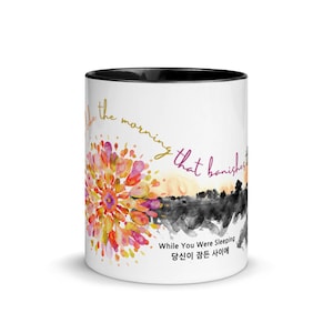 Puede incluir: Taza de cerámica blanca con borde negro. La taza presenta un diseño de acuarela de una puesta de sol con una flor en primer plano. El texto "like the morning that banishes" está escrito en letras doradas alrededor de la flor. El texto "While You Were Sleeping 당신이 잠든 사이에" está escrito en letras negras en la parte inferior de la taza.