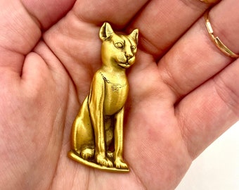 Op de afbeelding: Een gouden metalen beeldje van een zittende kat. De kat heeft een gedetailleerd gezicht en een halsband met een klein hangertje.