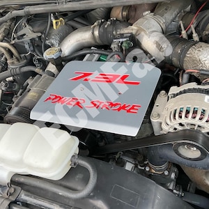 May include: A close-up of a grey engine compartment with a silver engine cover. The cover has red lettering that reads "7.3L POWER STROKE".