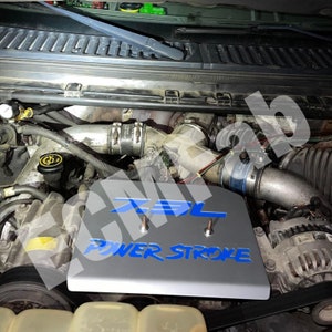 May include: A silver engine cover with a blue "7.3L POWER STROKE" logo. The cover is installed on a vehicle engine.