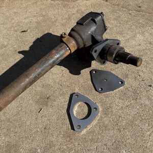 May include: An automotive steering gear assembly with a rusty, cylindrical shaft and a black, rectangular housing. Two black metal mounting plates are also present. The parts are arranged on a concrete surface.