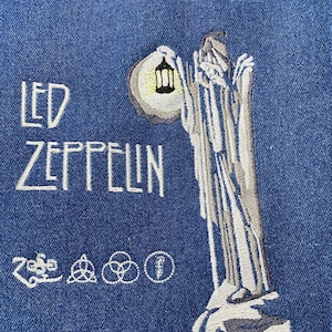 Puede incluir: Una chaqueta vaquera azul con texto bordado en blanco que dice "LED ZEPPELIN". El bordado también incluye una figura sosteniendo una linterna, una luna y cuatro símbolos.