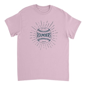 May include: A light pink short-sleeve t-shirt featuring a dark blue graphic on the chest. The graphic depicts a baseball with the text "HACKNEY" above, "ROUNDERS" in the center, and "DOWNS" below, all surrounded by radiating sunburst lines.