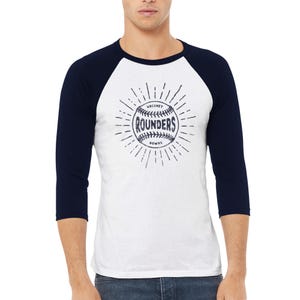 May include: A white baseball-style t-shirt with three-quarter length navy blue sleeves. The front features a dark blue graphic of a baseball with the text "HACKNEY ROUNDERS" printed in the center, surrounded by radiating sunburst lines.