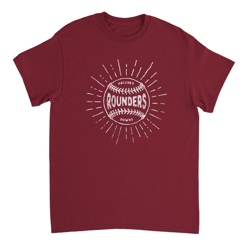May include: A maroon short-sleeved t-shirt featuring a white graphic of a baseball with the text "HACKNEY ROUNDERS DOWNS" printed across the center. The design includes radiating lines around the ball, creating a sunburst effect on the chest.