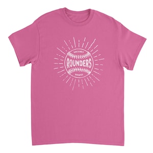 May include: A pink short-sleeved t-shirt featuring a white graphic of a baseball with the text "HACKNEY ROUNDERS" printed in the center. The design includes radiating lines extending outward from the ball, creating a sunburst effect on the chest.