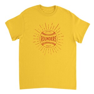 May include: A mustard yellow short-sleeve t-shirt featuring a central graphic of a baseball with red stitching. The text "HACKNEY ROUNDERS DOWNS" is printed on the ball, surrounded by radiating sunburst lines in a matching red color.