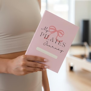 K&ouml;nnte beinhalten: Ein hellrosa Notizbuch mit einer zartrosa Schleifenillustration und dem schwarzen Text "My PILATES Journey" auf dem Einband. Unter dem Text befindet sich ein leeres wei&szlig;es rechteckiges Feld zum Schreiben. Der Einband enth&auml;lt auch "DIGITAL DOWNLOAD".