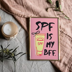 May include: A framed pink print with the text "SPF is my BFF."  A yellow tube of sunscreen with a sun graphic and the word "SPF" is pictured.