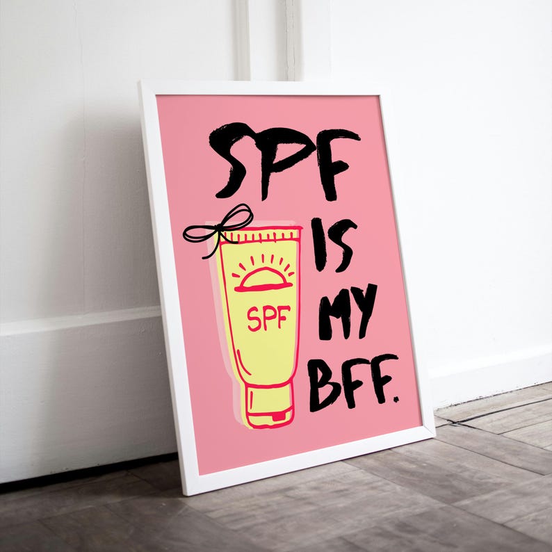 May include: A pink framed print with a white background and black text that reads "SPF is my BFF." A yellow tube of sunscreen with a red sun and the word "SPF" is pictured in the centre of the print.