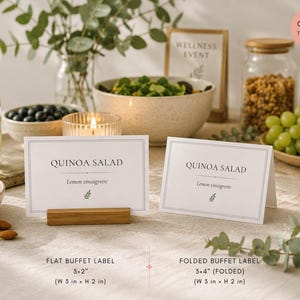 May include: Two white rectangular buffet labels with black text reading QUINOA SALAD and Lemon vinaigrette, featuring a small green leaf icon. One label is flat on a wooden stand, the other is folded. Dimensions are 3x2 inches and 3x4 inches.