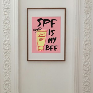 May include: A pink framed print with a white background and black text that reads "SPF is my BFF." A cartoon image of a yellow sunscreen bottle with a red sun and the word "SPF" is on the left side of the text.