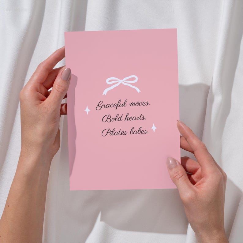 Puede incluir: Una tarjeta rosa con una ilustraci&oacute;n de lazo blanco y el texto "Graceful moves. Bold hearts. Pilates babes." La tarjeta es sostenida por dos manos sobre un fondo blanco.