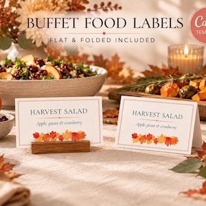 May include: Two white rectangular buffet food labels with a thin border and a row of colorful autumn leaves at the bottom. The text reads "HARVEST SALAD" and "Apple, pecan & cranberry". One label is flat in a wooden holder, the other is folded.