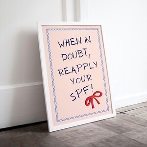 May include: A white framed print with a pink background and blue border. The print says "When in doubt, reapply your SPF!" with a red bow underneath.