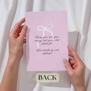 Puede incluir: Una tarjeta rosa claro con texto blanco y una ilustraci&oacute;n de lazo es sostenida por manos. La tarjeta dice: "Bring your mat, your energy, and your cutest pilates fit! Let's stretch, sip, and celebrate!" La palabra "BACK" est&aacute; abajo.