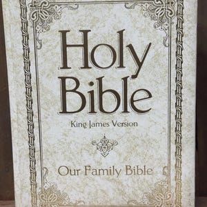 May include: A hardcover Holy Bible, King James Version, with the title in large, dark brown letters. The cover is off-white with a textured pattern and decorative gold-colored borders and accents. The words "Our Family Bible" are printed at the bottom.
