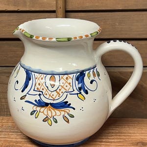 May include: A white ceramic pitcher with a rounded body and a curved handle. The pitcher is decorated with blue, orange, and yellow floral patterns. The rim is trimmed with green and orange accents. A decorative piece for the kitchen.