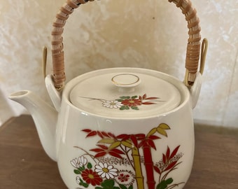 May include: White ceramic teapot with a bamboo and floral design. The teapot has a woven brown handle and a gold trim around the lid.