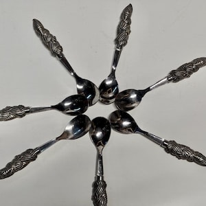 May include: A collection of seven silver spoons arranged in a circular pattern. Each spoon features a decorative handle with a detailed, textured design. The spoons are made of a shiny, reflective metal.