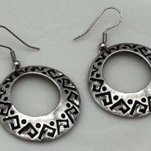 May include: A pair of silver-toned hoop earrings with a decorative black geometric pattern around the outer edge. The earrings have a hook closure and are displayed on a light-colored surface.